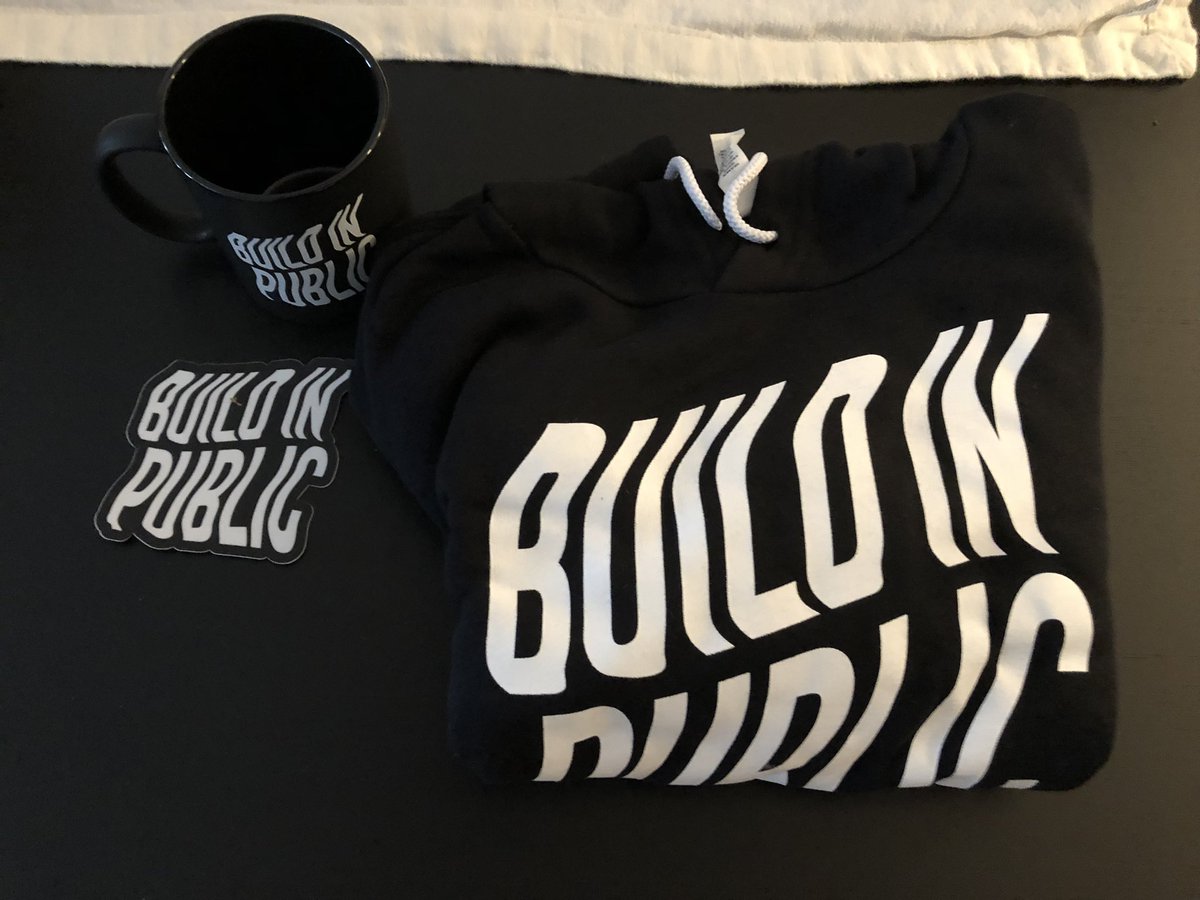 A picture of a folded up sweatshirt, a coffee mug and a sticker that all say “Build In Public”.