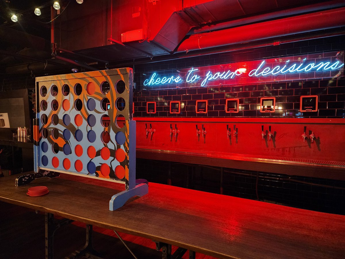 A huge game of Connect 4 sitting on a table with a neon sign on the back wall saying "cheers to pour decisions" above a row of beer taps.
